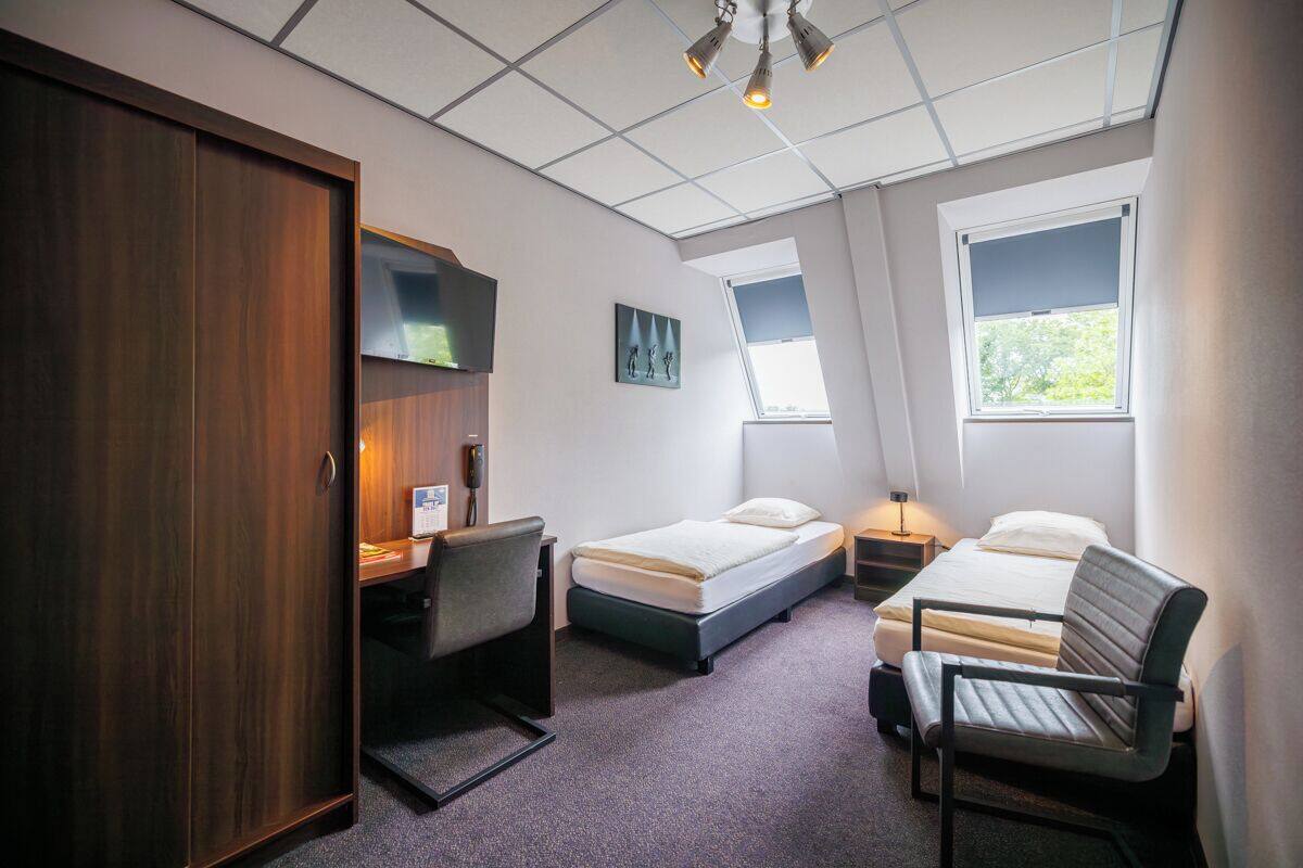 A compact hotel room with a desk, two single beds, a wardrobe, and a large window providing natural light.