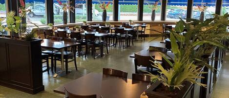 Un restaurant au décor moderne, avec un mélange de banquettes et de tables, de grandes fenêtres donnant sur le parking et des plantes en pot qui apportent une touche de verdure.