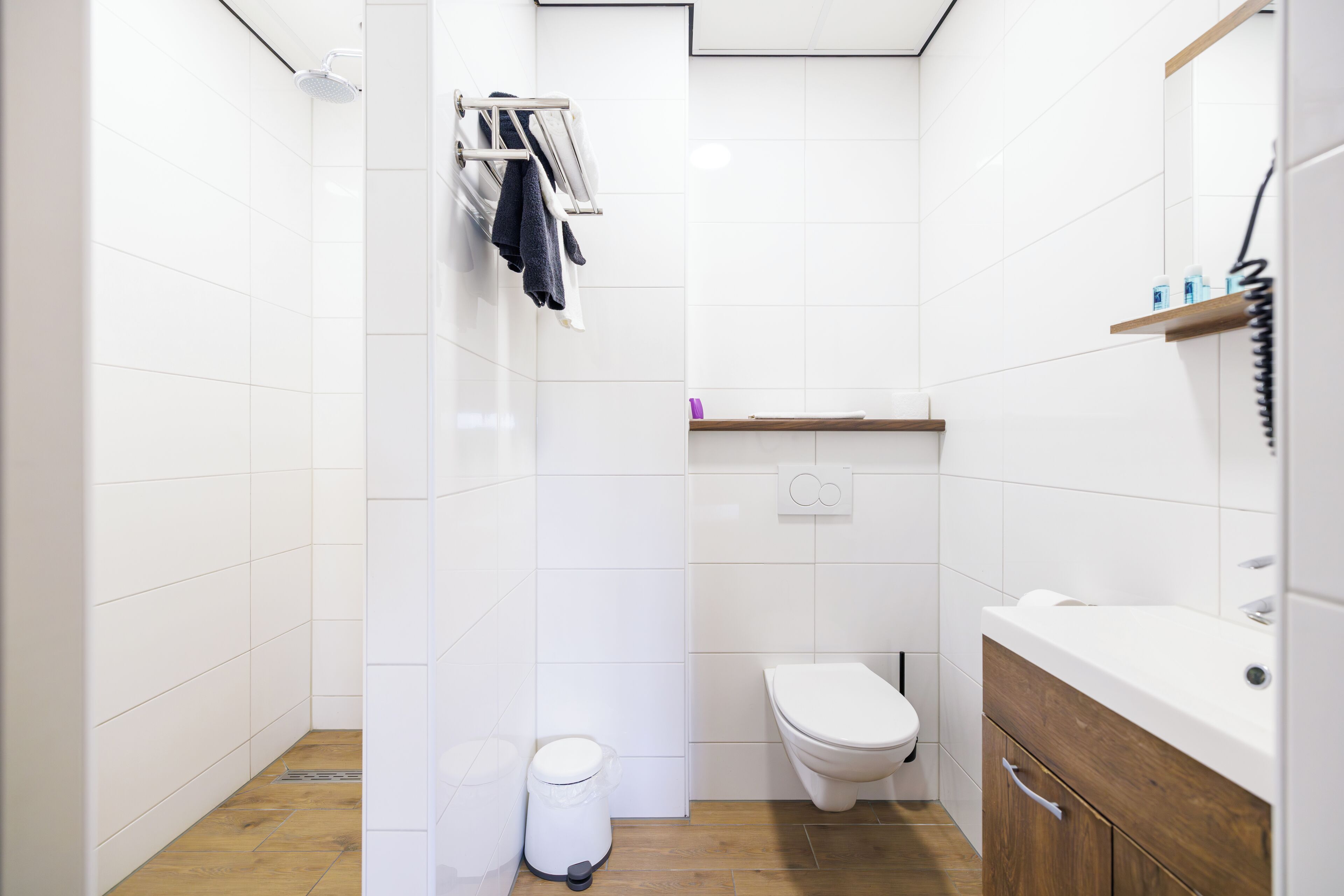 Un bagno moderno con piastrelle bianche, pavimento in legno, un water bianco, una doccia e un mobile bagno in legno con uno specchio.