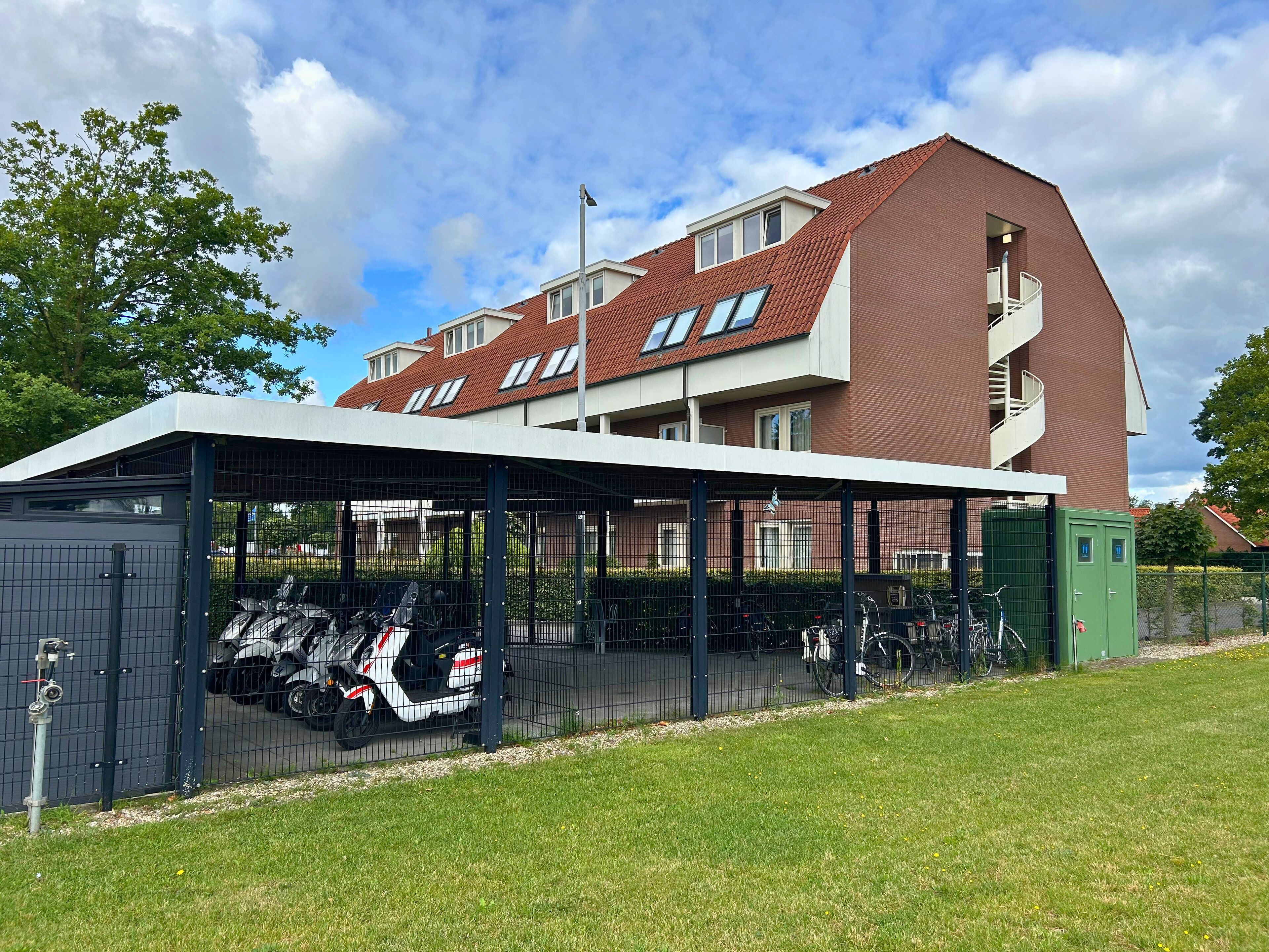 A modern building with a covered parking area, multiple windows, and a green fence.