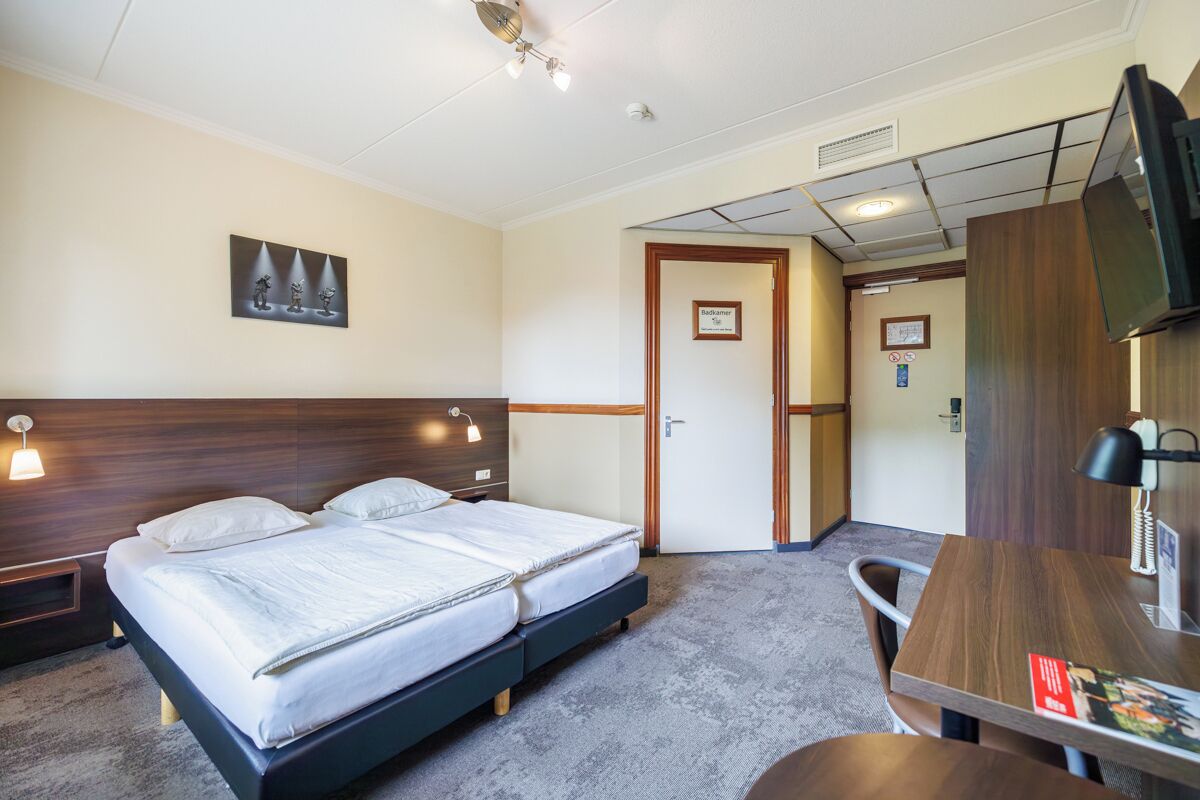 A hotel room with a double bed, a desk with a chair, a TV, and a door leading to a bathroom.