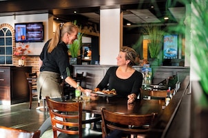 Un interior de restaurante con mesas y sillas de madera, una zona de bar y una pantalla de televisión mostrando un menú.