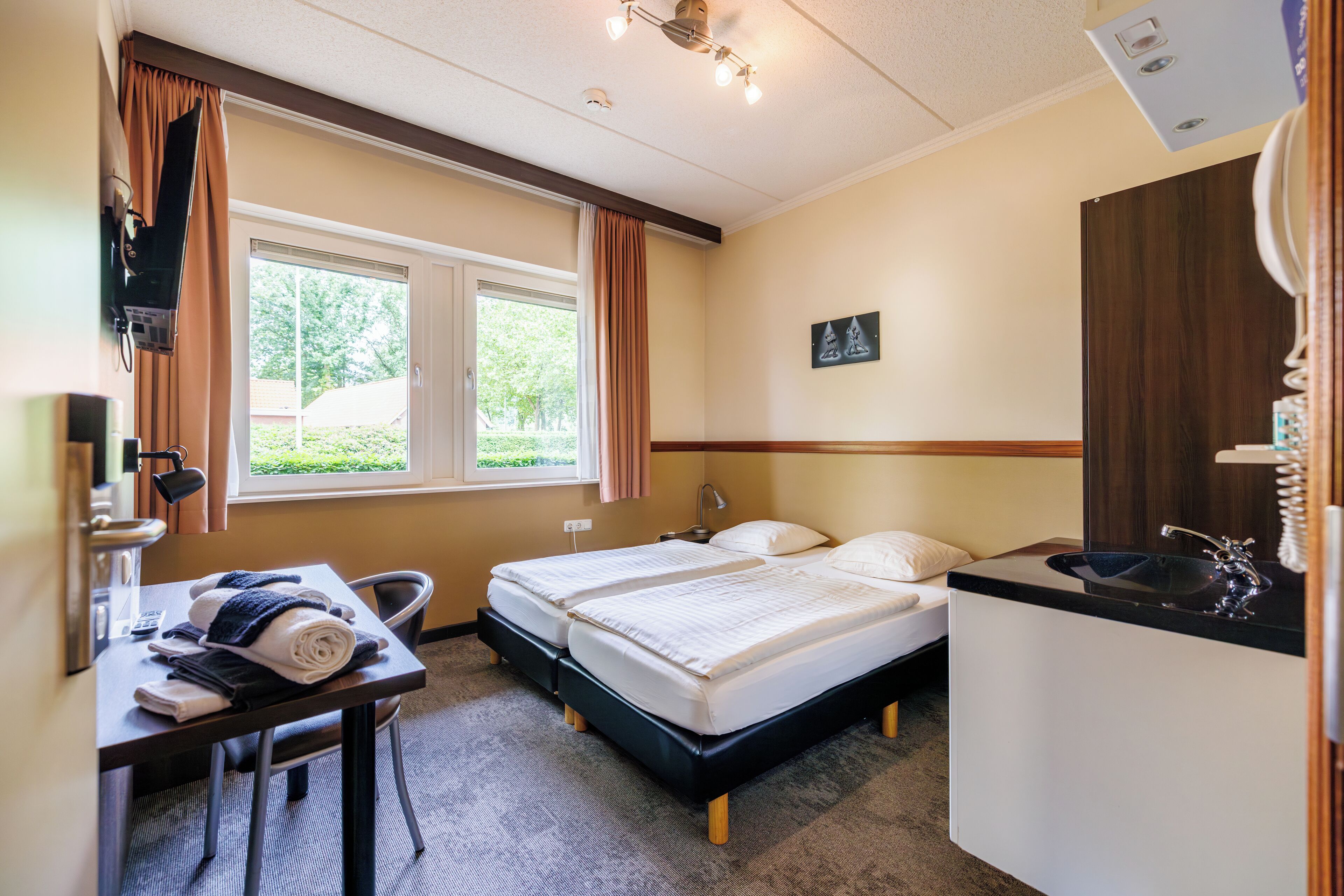 A hotel room with two beds, a desk, a chair, a window with curtains, a sink, and a mirror.
