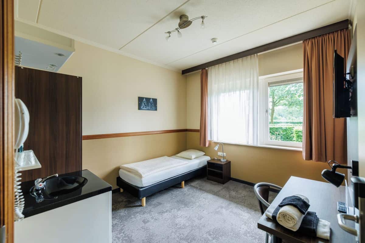 A compact hotel room with a single bed, a small desk with a chair, a bedside table, a mirror, a window with curtains, and a television.