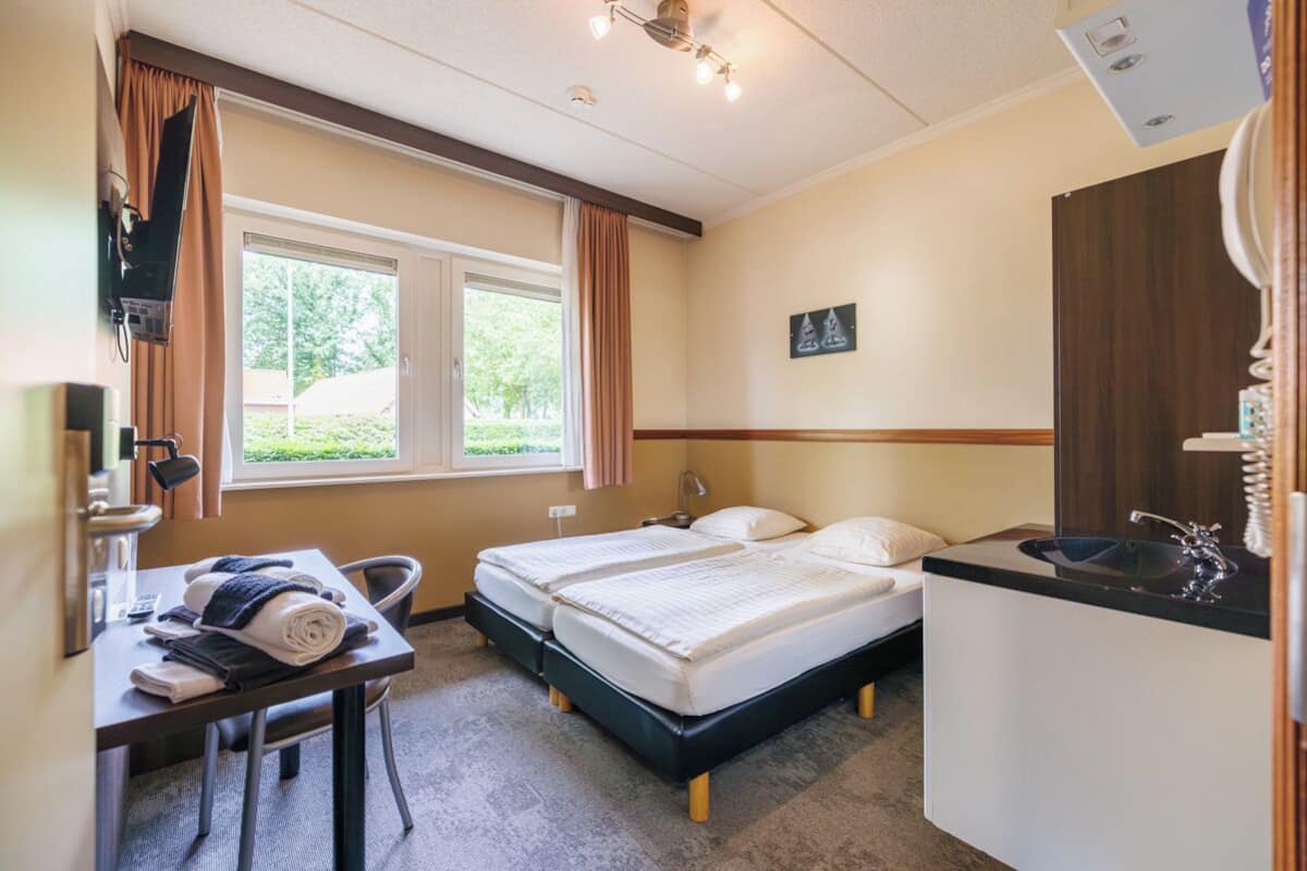 A hotel room with a bed, a desk with a chair, a sink, and a window with curtains.