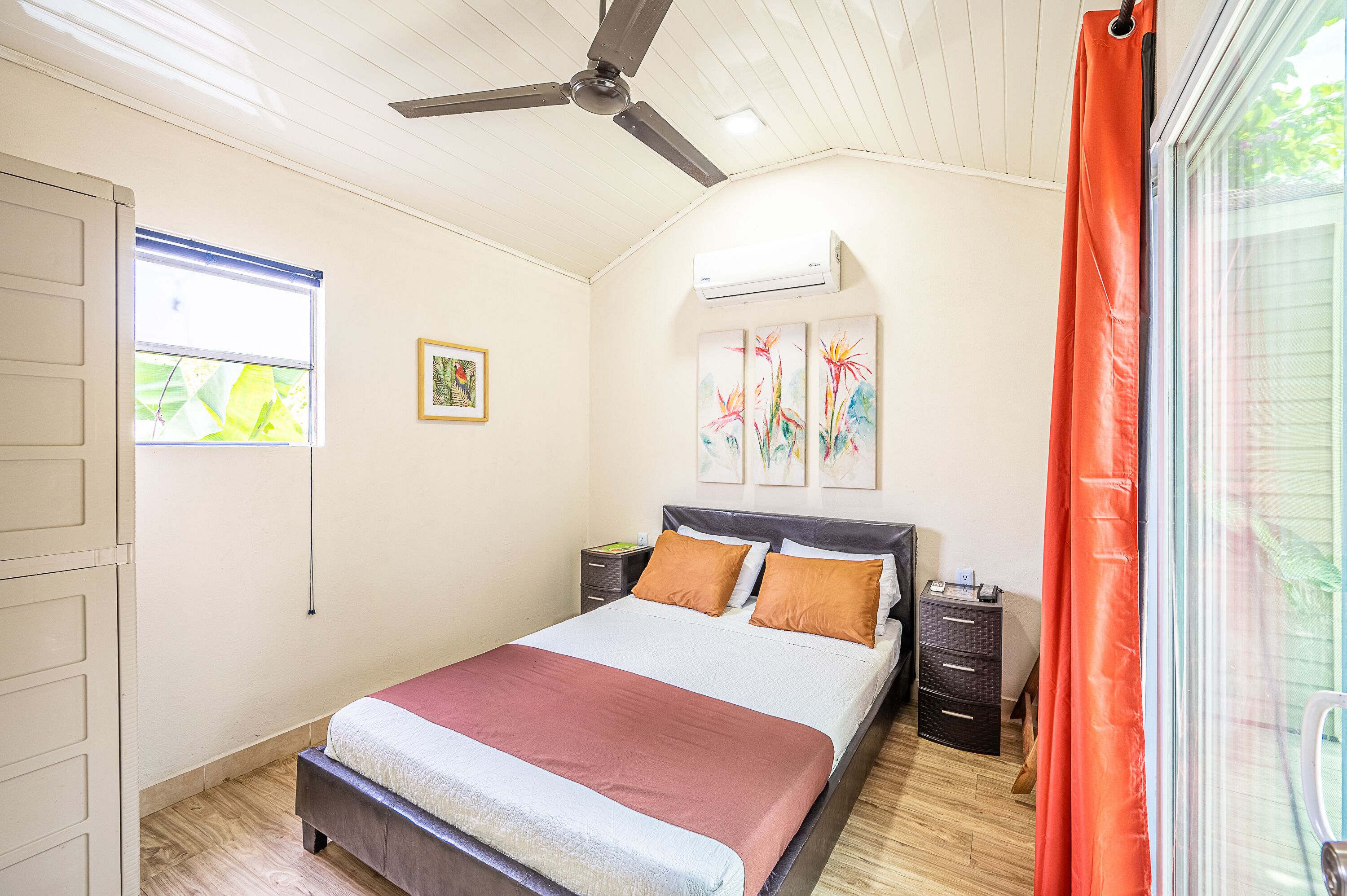 A bedroom with a bed, a ceiling fan, an air conditioning unit, a window with blinds, a nightstand, and a drawer unit.