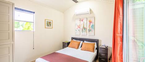 A bedroom with a bed, a ceiling fan, an air conditioning unit, a window with blinds, a nightstand, and a drawer unit.