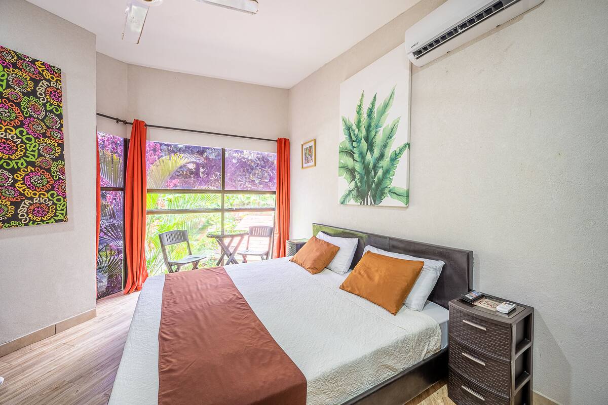 A bedroom with a large bed, a window with curtains, a chair, and a plant on the wall.