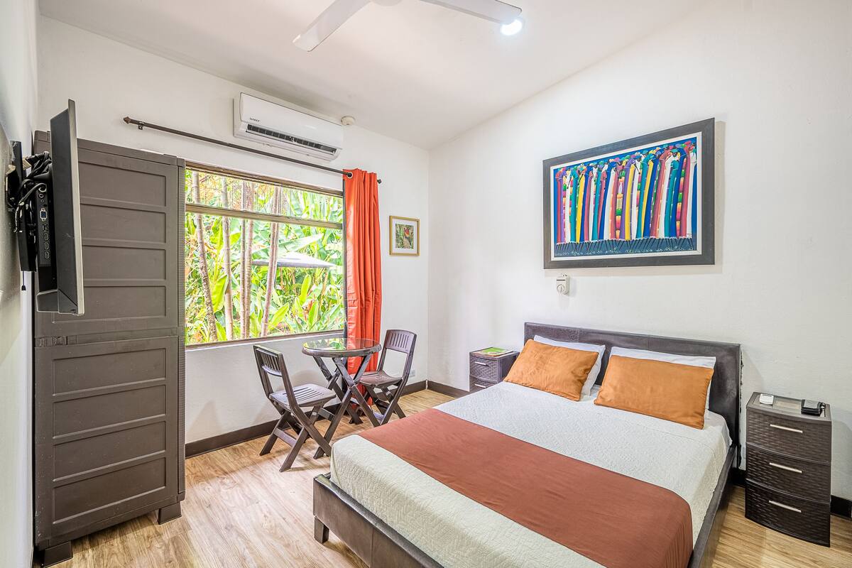 A bedroom with a bed, a desk, and a chair. There is a window with curtains, a picture on the wall, and a small table.