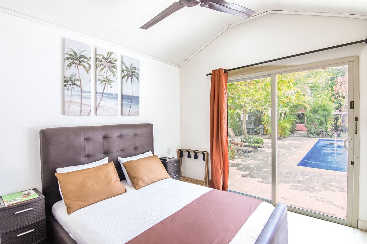 A bedroom with a bed, a ceiling fan, a nightstand, and a view of an outdoor swimming pool.