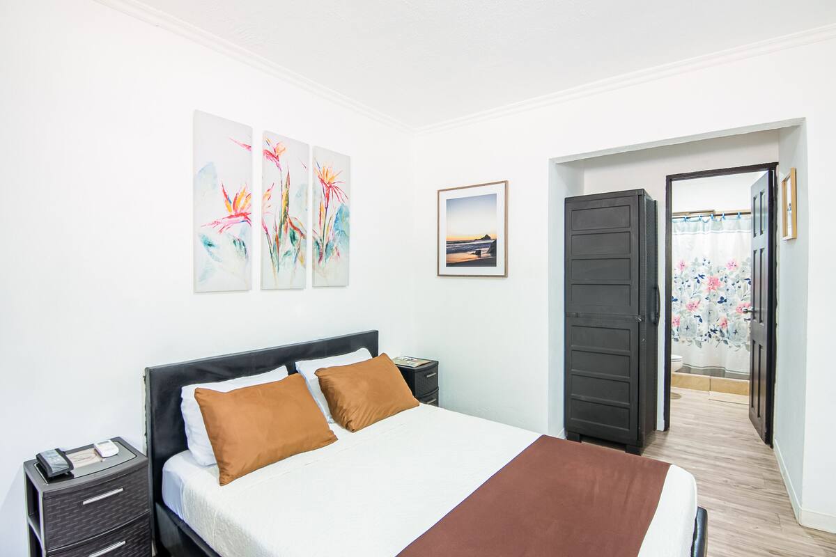 A hotel room with a bed, a nightstand, a framed picture, and a door leading to a bathroom.