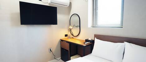 A hotel room with a bed, a desk with a mirror, a flat-screen TV, an air conditioner, and a window with blinds.
