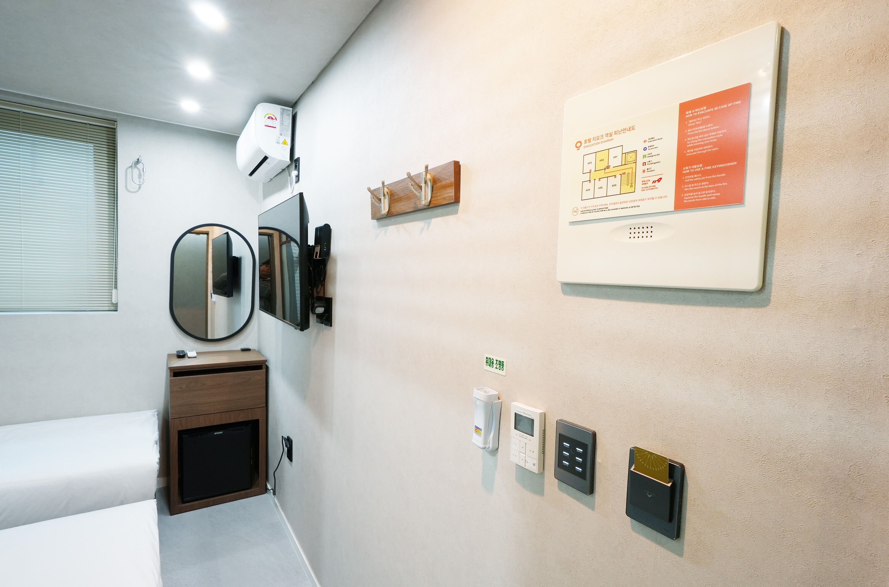 A hotel room with a bed, a small bedside table, a mirror, a wall-mounted air conditioner, a wall-mounted thermostat, a smoke detector, and a fire alarm.