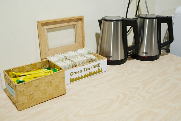 Una caja de madera con la etiqueta 'Té Verde' contiene bolsitas de té, dos teteras de acero inoxidable y una tetera eléctrica negra sobre una superficie de madera.