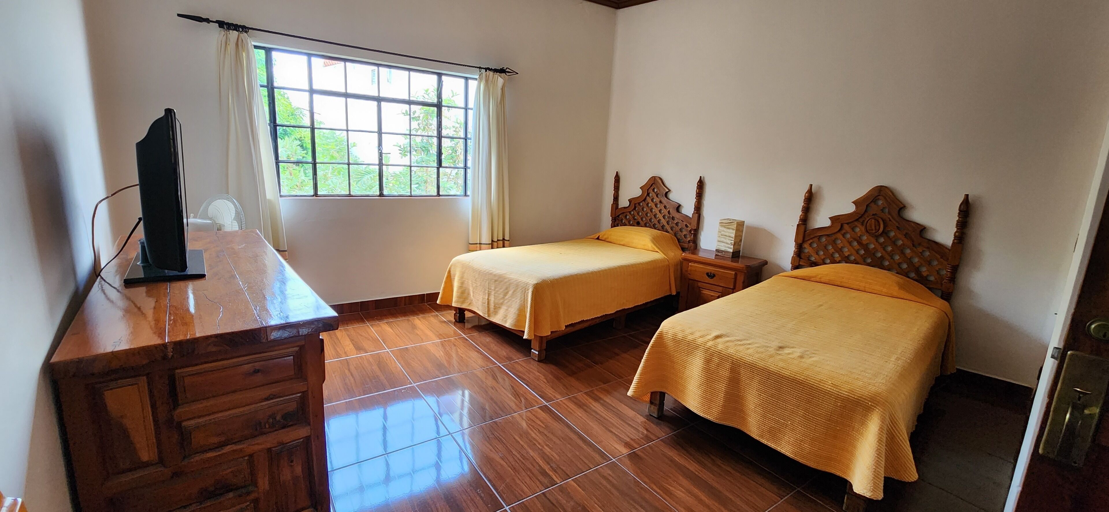 Une chambre avec deux lits, un bureau en bois, une chaise, une fenêtre avec des rideaux et un sol carrelé.