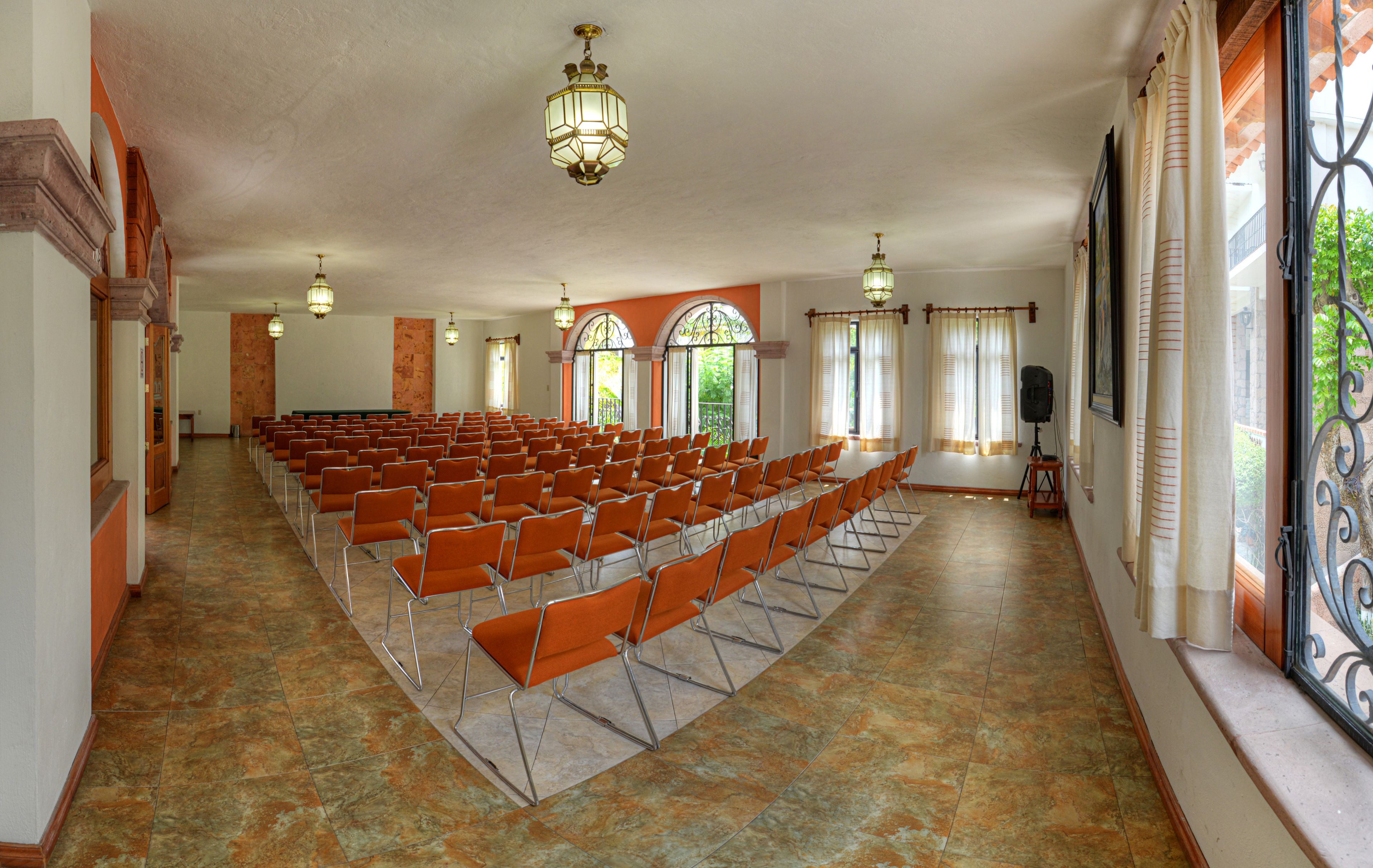 A spacious hall with rows of orange chairs, large windows with curtains, and a speaker on a stand.