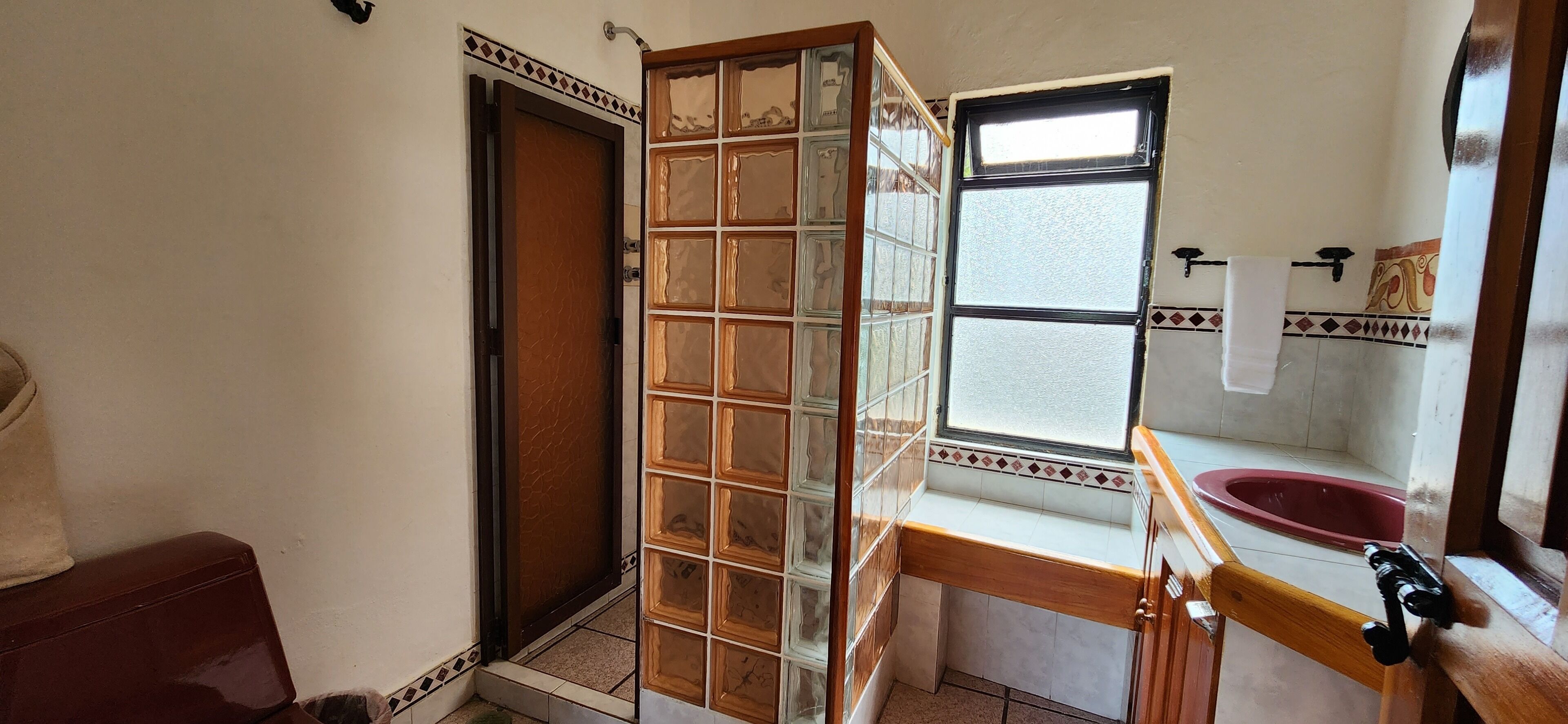 A bathroom with a wooden door, a window with a view, a red sink, and a tiled floor.