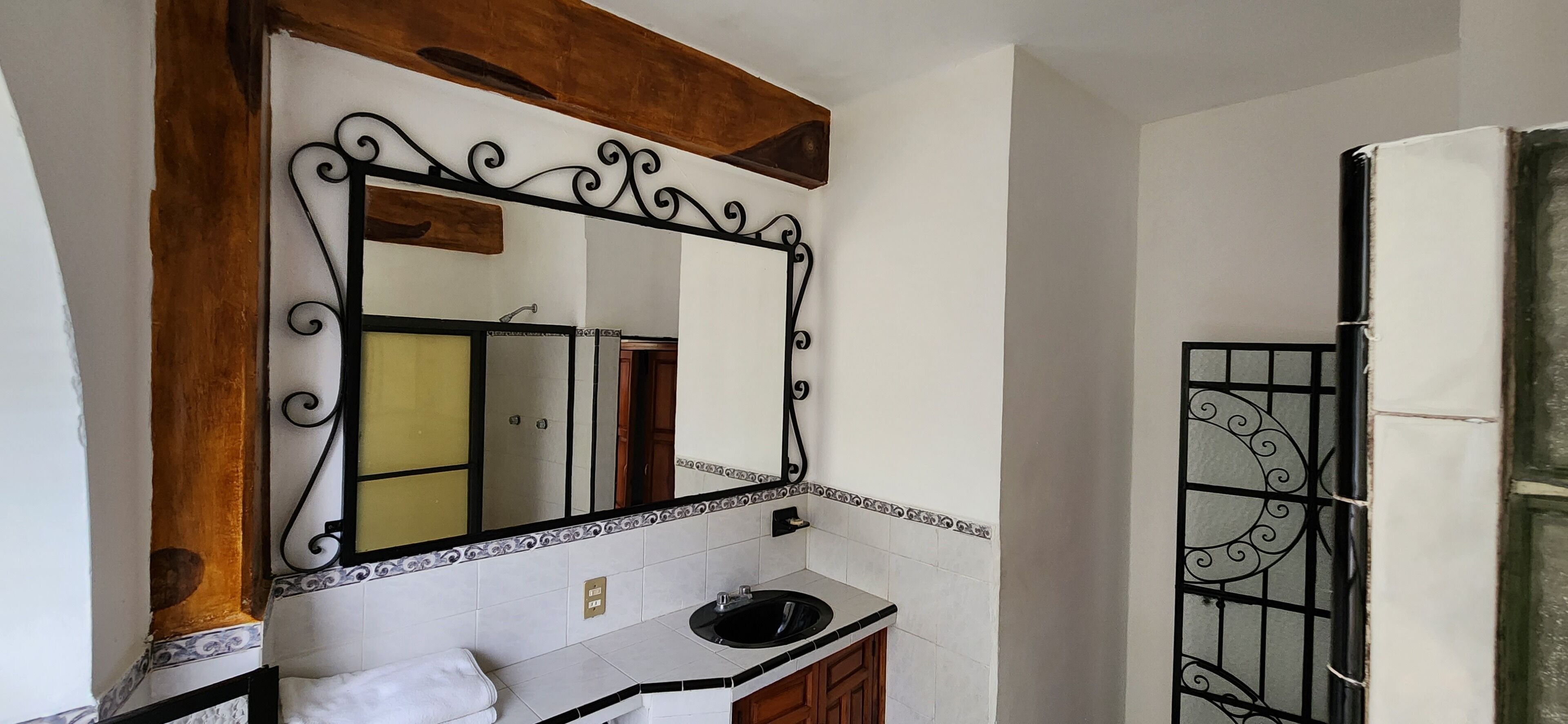 Une salle de bain avec un lavabo blanc, un miroir doté d’un cadre décoratif et un espace douche séparé par une porte en métal.