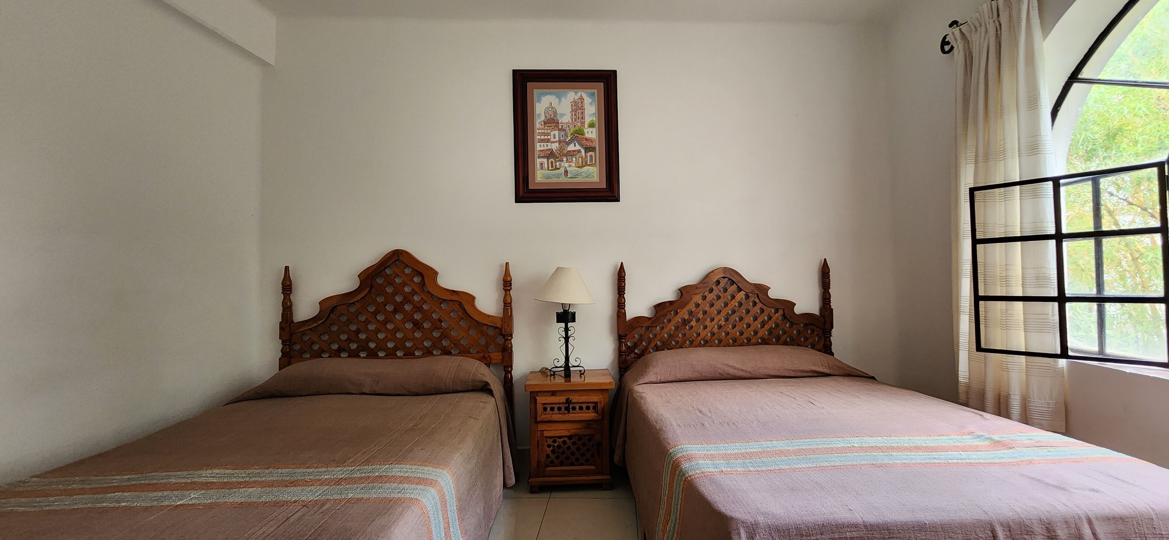 Una habitación con dos camas de madera, una mesita de noche con lámpara, un cuadro en la pared y una ventana con cortinas.