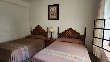 A room with two single beds, a nightstand with a lamp, a framed picture on the wall, and a window with a view.