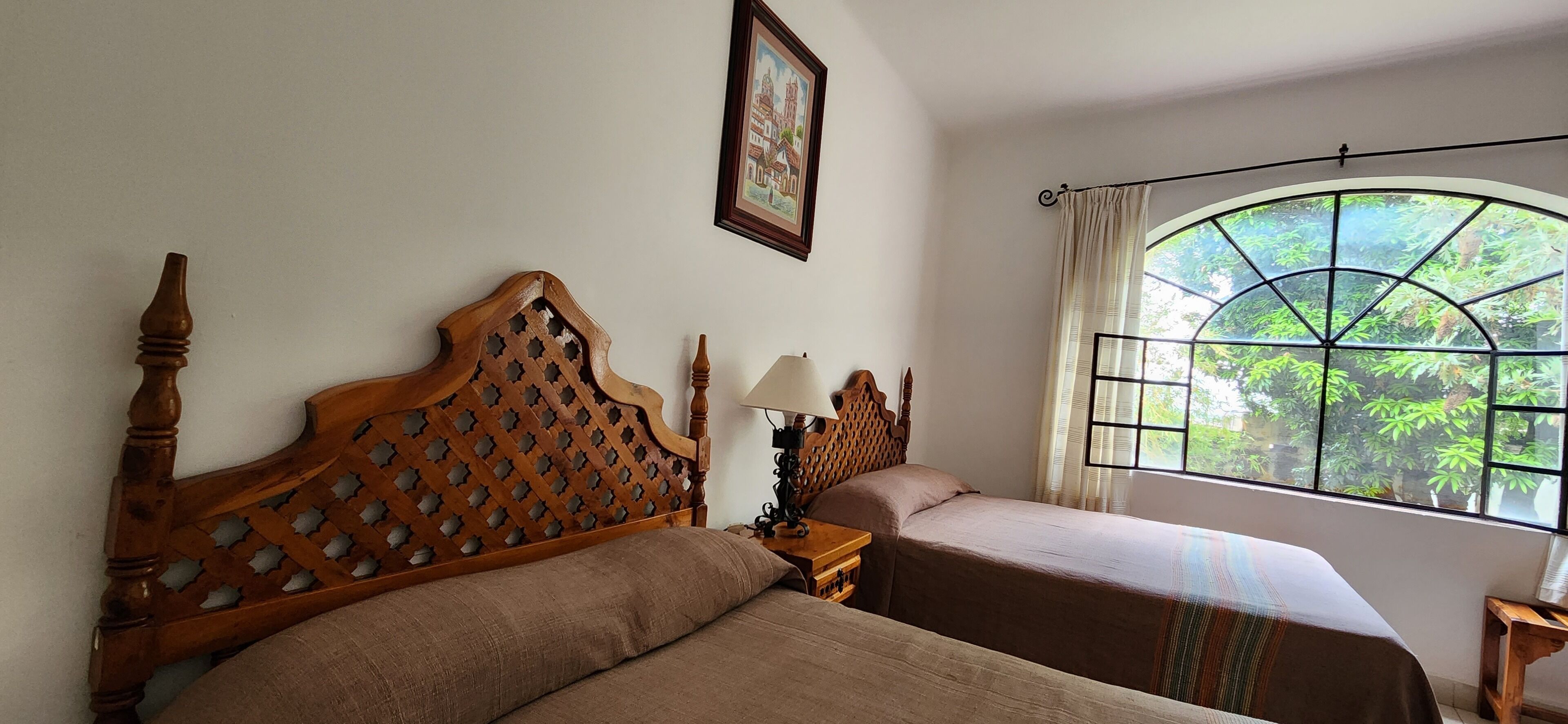 A room with two beds, a wooden headboard, a nightstand with a lamp, and a window with a view of greenery.