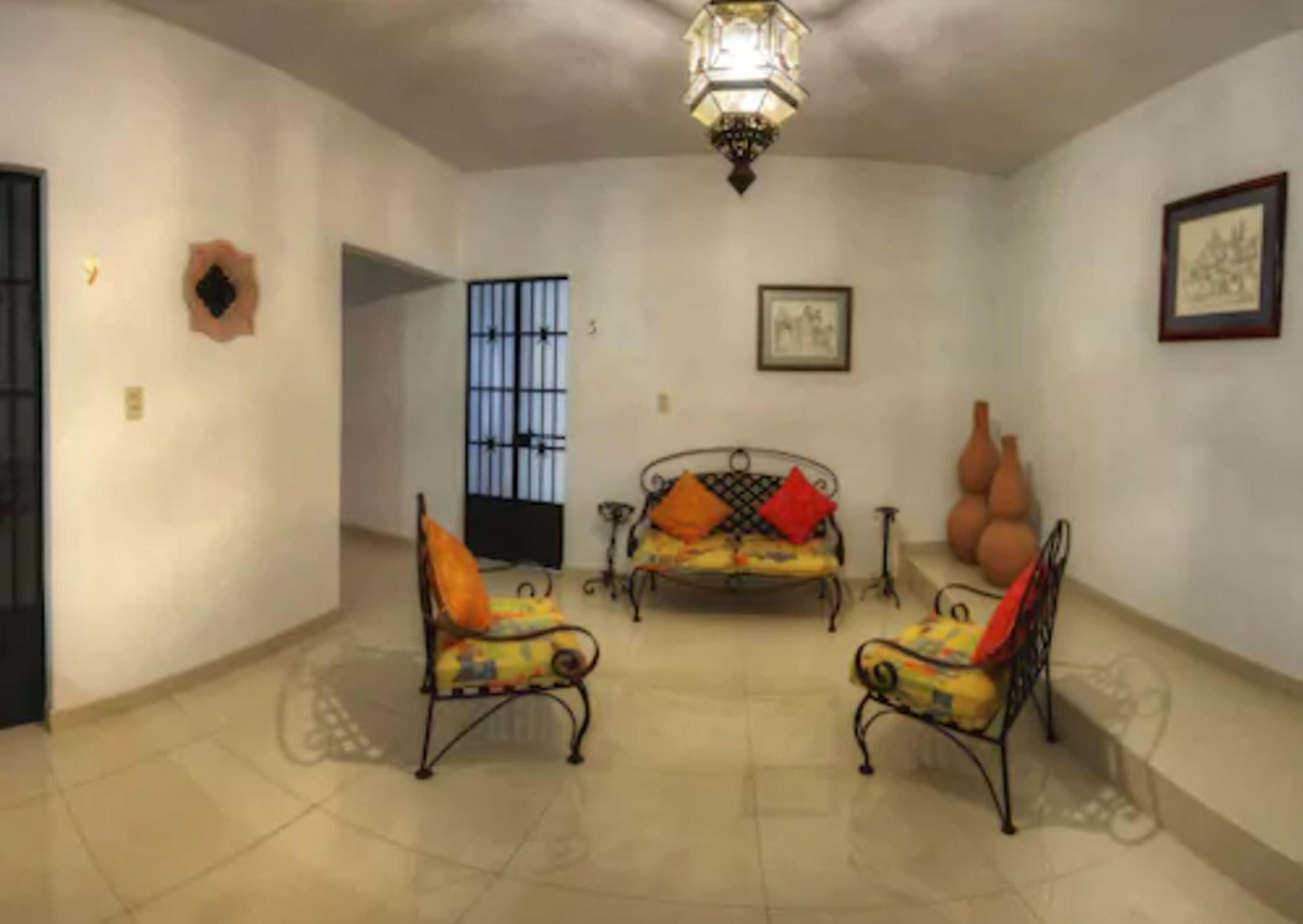 A spacious room with a high ceiling, a chandelier, a sofa with cushions, two armchairs, a large window with bars, and a wall-mounted picture.