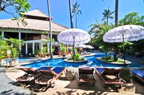 A resort pool area with lounge chairs and umbrellas, surrounded by palm trees and a building with a thatched roof. - Sativa Sanur Cottages (Denpasar)