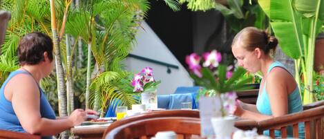 An outdoor dining area with wooden tables and chairs, surrounded by lush tropical plants and a building in the background.