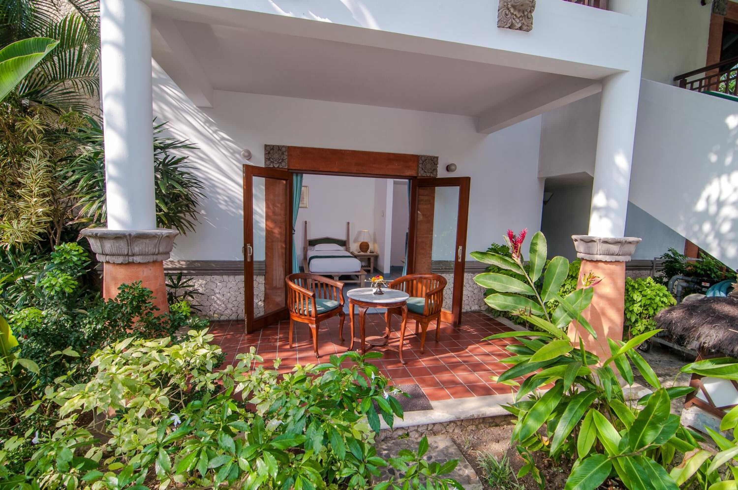 A covered outdoor seating area with a table and chairs, surrounded by lush greenery and a tiled floor.