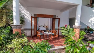 A covered outdoor seating area with a table and chairs, surrounded by lush greenery and a tiled floor.