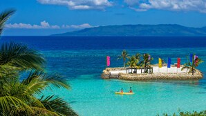 A tropical beach scene with clear turquoise waters, a small island with colorful flags, and a distant mountain range. - Mithi Resort & Spa (Dauis)