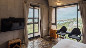 A hotel room with a large window offering a view of a coastal town, a flat-screen TV mounted on the wall, and two chairs near the window. - The Light Inn (Hengchun)