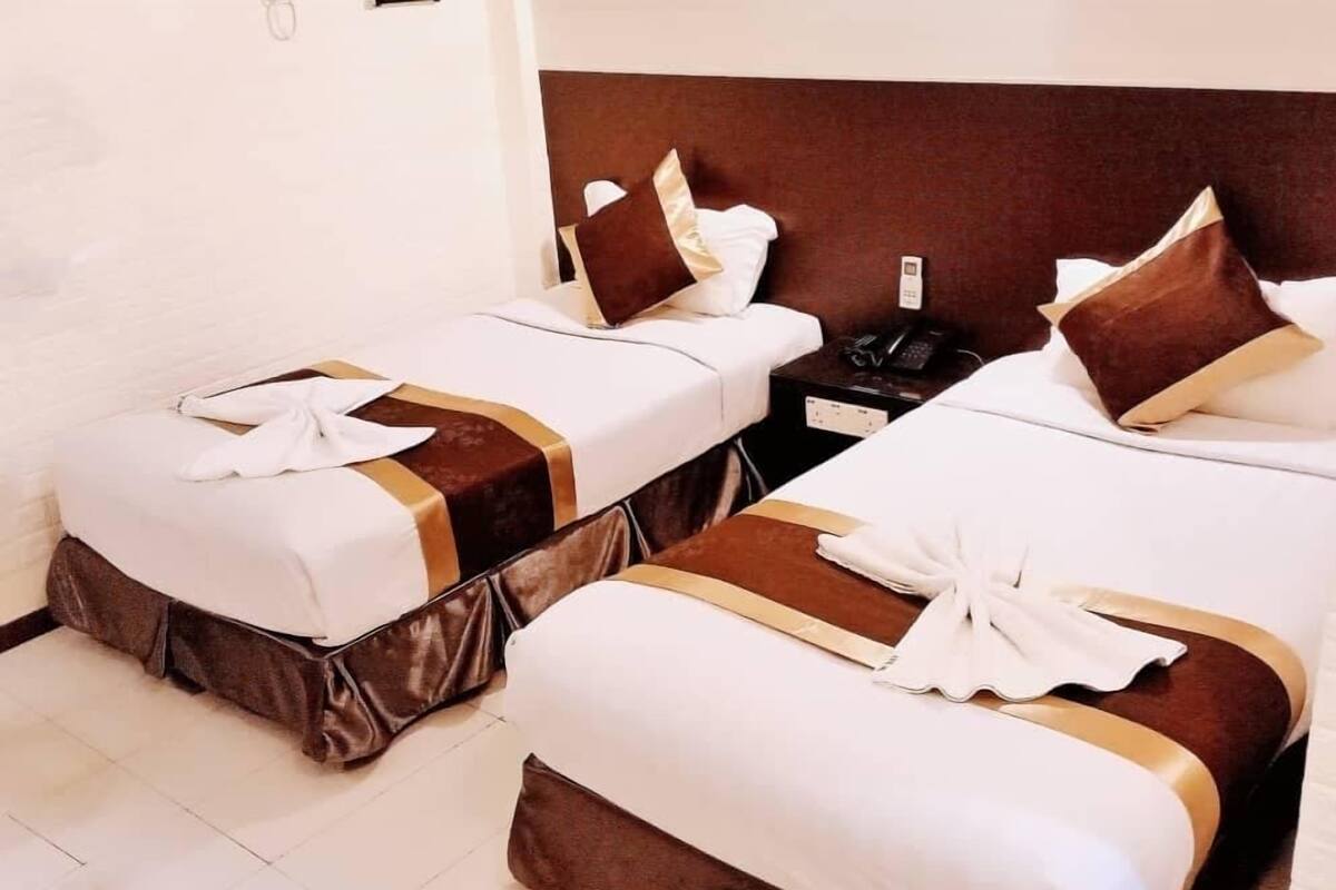 A hotel room with two single beds, each with a brown headboard, white bedding, and a white towel on the bed. There is a nightstand with a phone and a lamp between the beds.