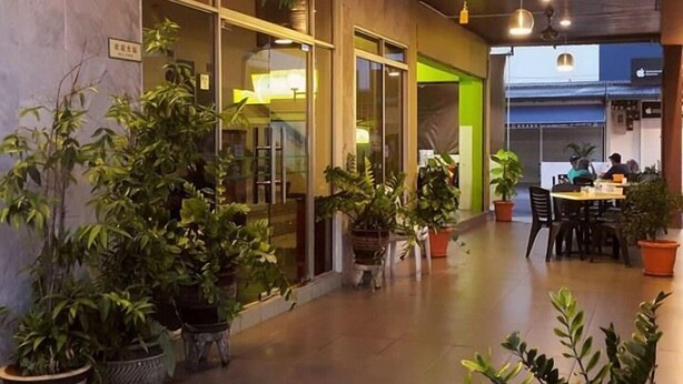 Un espacio interior con plantas en macetas, un área de estar con mesas y sillas, y un acceso por puerta de vidrio.