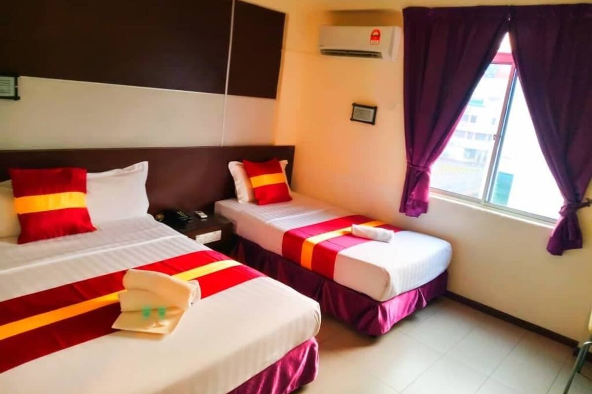 A hotel room with two beds, each with red and white bedding, a nightstand, an air conditioning unit, and a window with purple curtains.