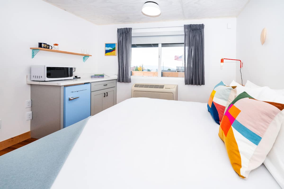 A hotel room with a bed, a desk with a microwave, a small cabinet, a window with curtains, and a flag outside.