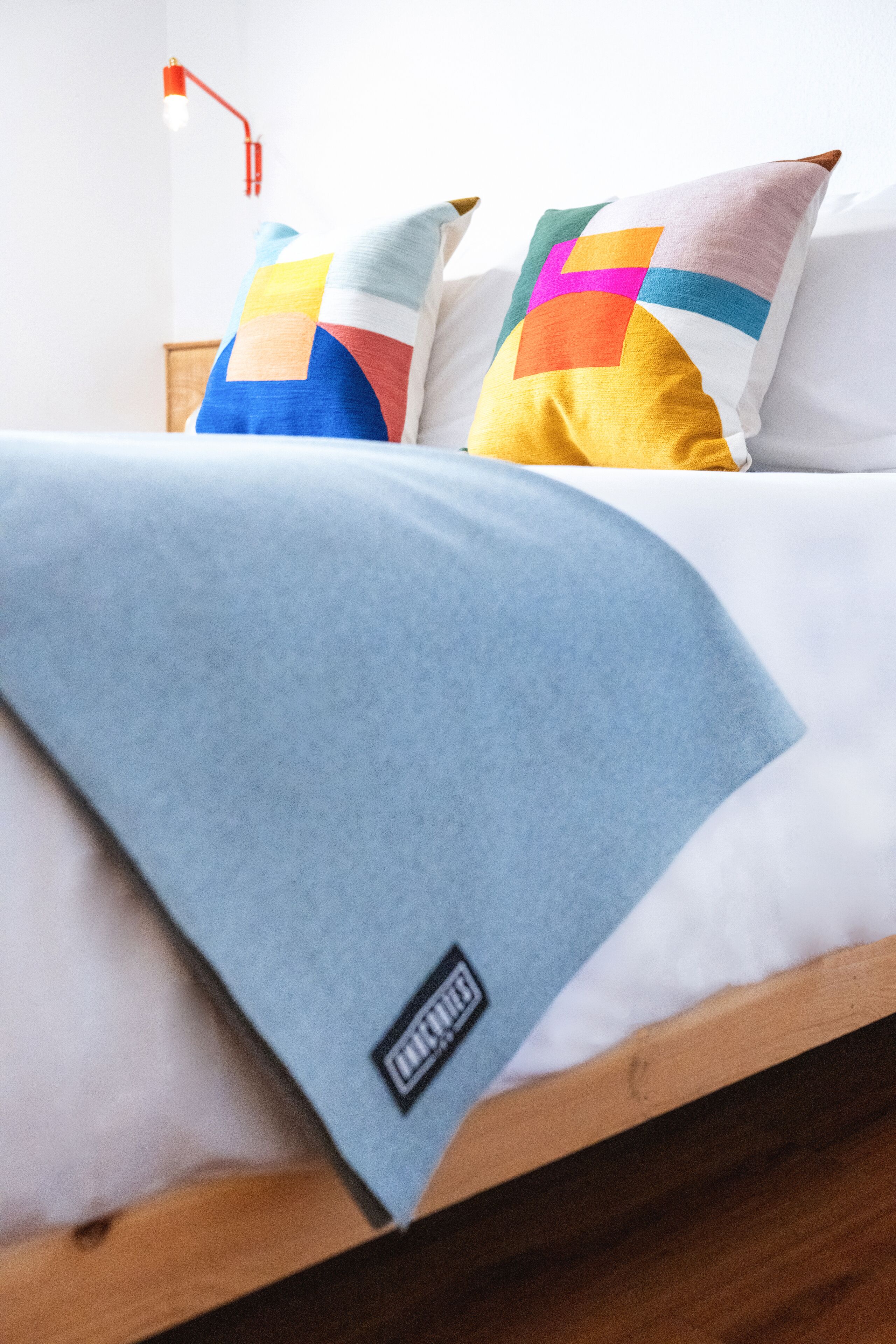 Ein Bett mit einer blauen Decke, zwei Kissen mit bunten Mustern und einer roten Lampe auf einem Nachttisch aus Holz.