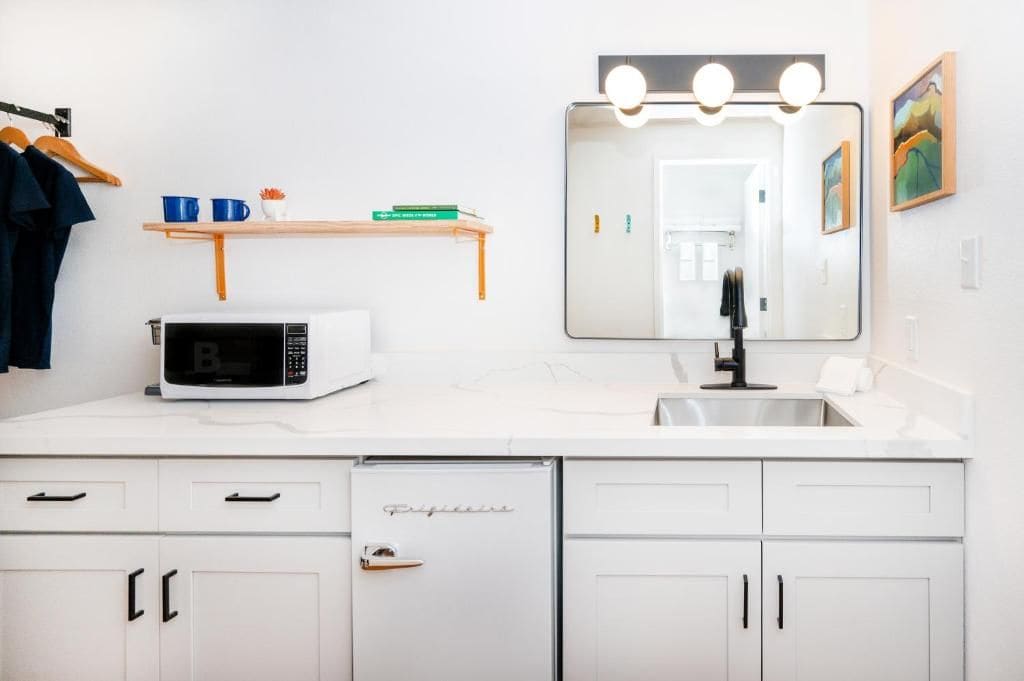 A modern bathroom with a white countertop, a microwave, a sink with a mirror above, and a shelf with items.