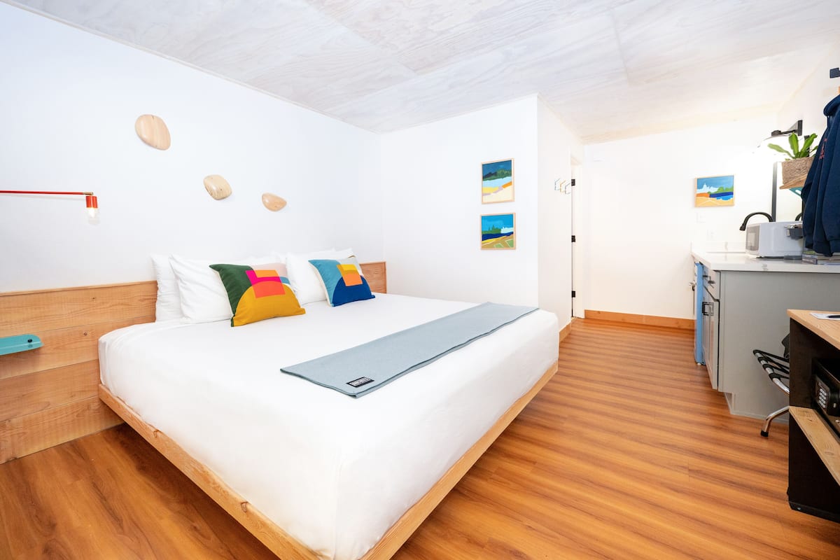 A modern hotel room with a wooden floor, a bed with colorful pillows, a desk with a plant, and artwork on the walls.