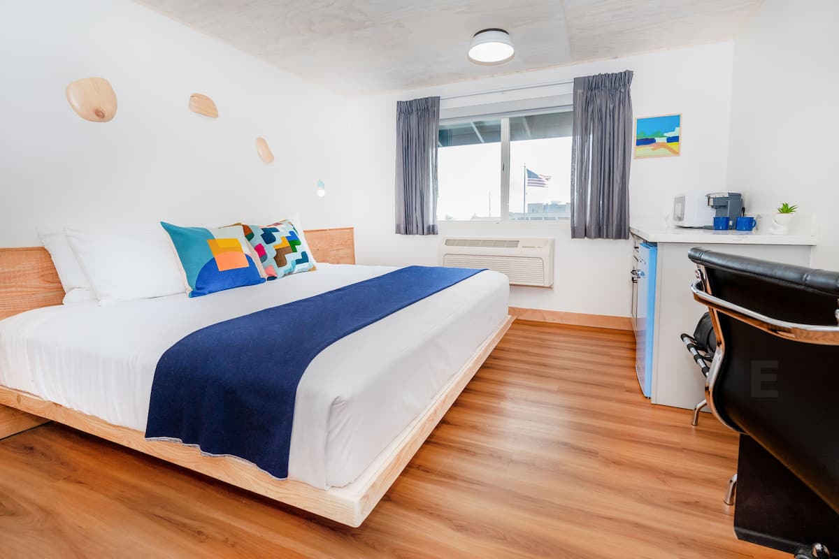 A modern hotel room with a wooden floor, a bed with a blue blanket, a desk with a coffee maker, and a view of the city skyline.