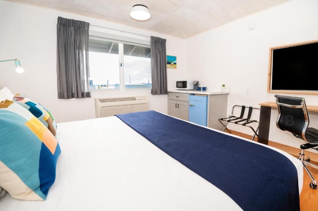 A hotel room with a bed, a desk, a chair, a microwave, a small cabinet, and a window with curtains.