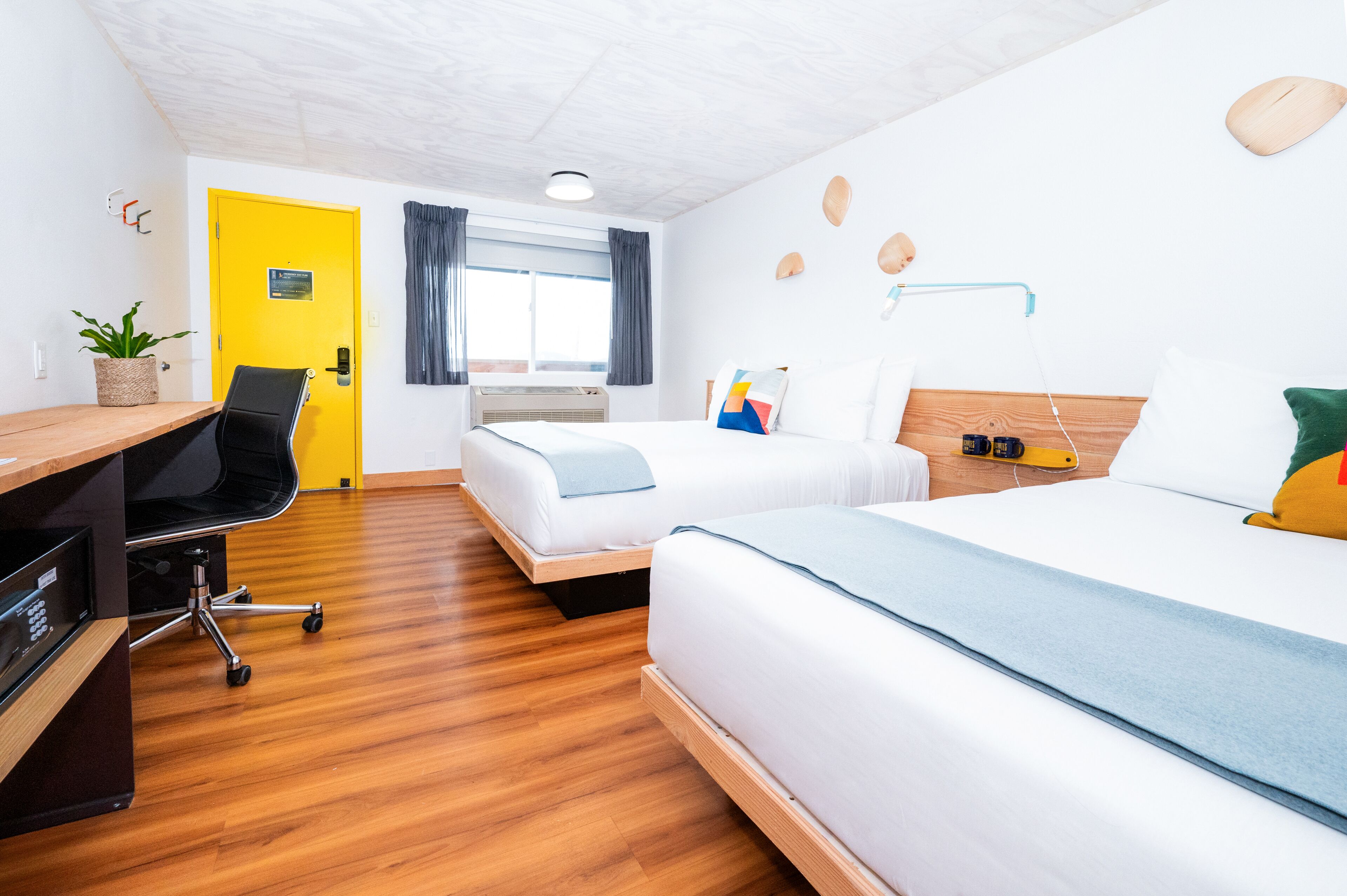 A hotel room with a wooden floor, a desk with a chair, two beds, a small table, and a window with curtains.
