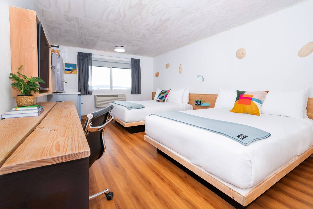 A modern hotel room with two beds, a wooden desk, a chair, a potted plant, and a window with curtains.