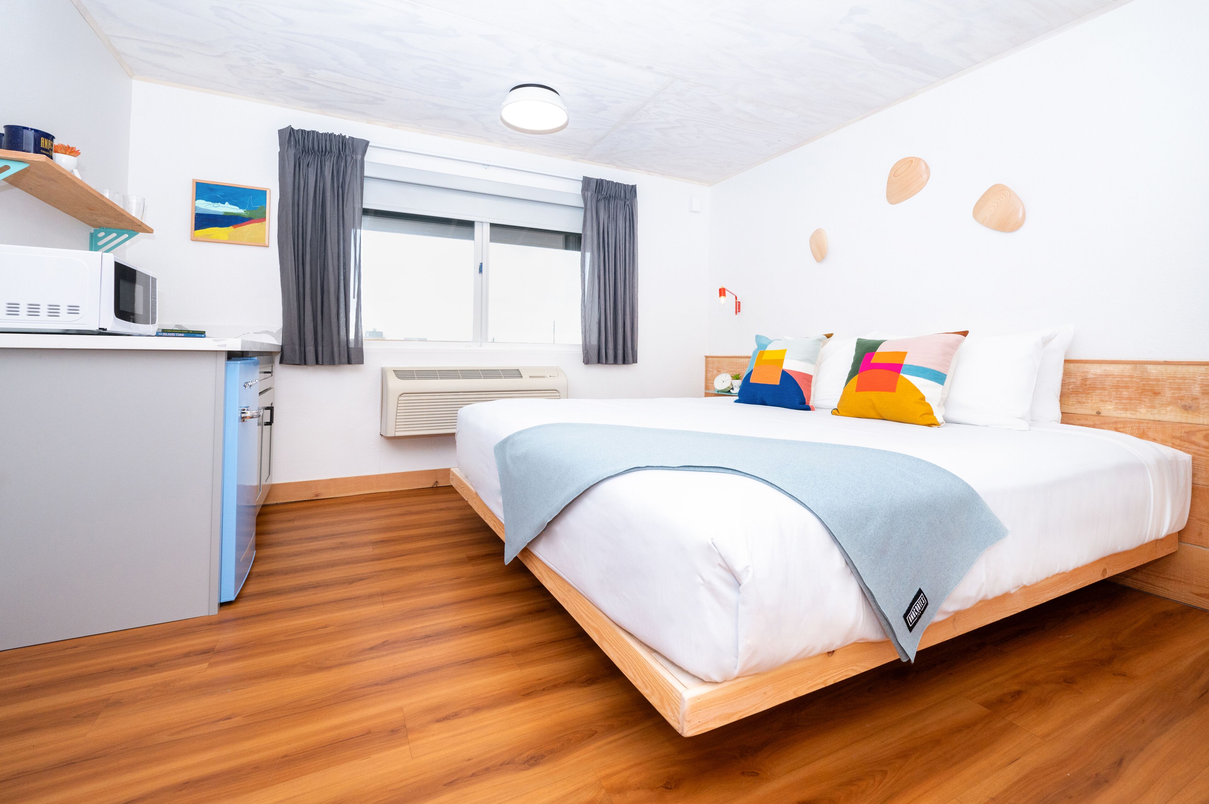 Una habitación de hotel moderna con suelo de madera, una cama con almohadas de colores, un escritorio con microondas y una ventana con cortinas.