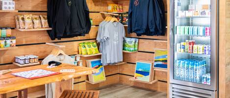 A room with wooden walls, a wooden table, a bench, a refrigerator stocked with beverages, and various food items and clothing on shelves.