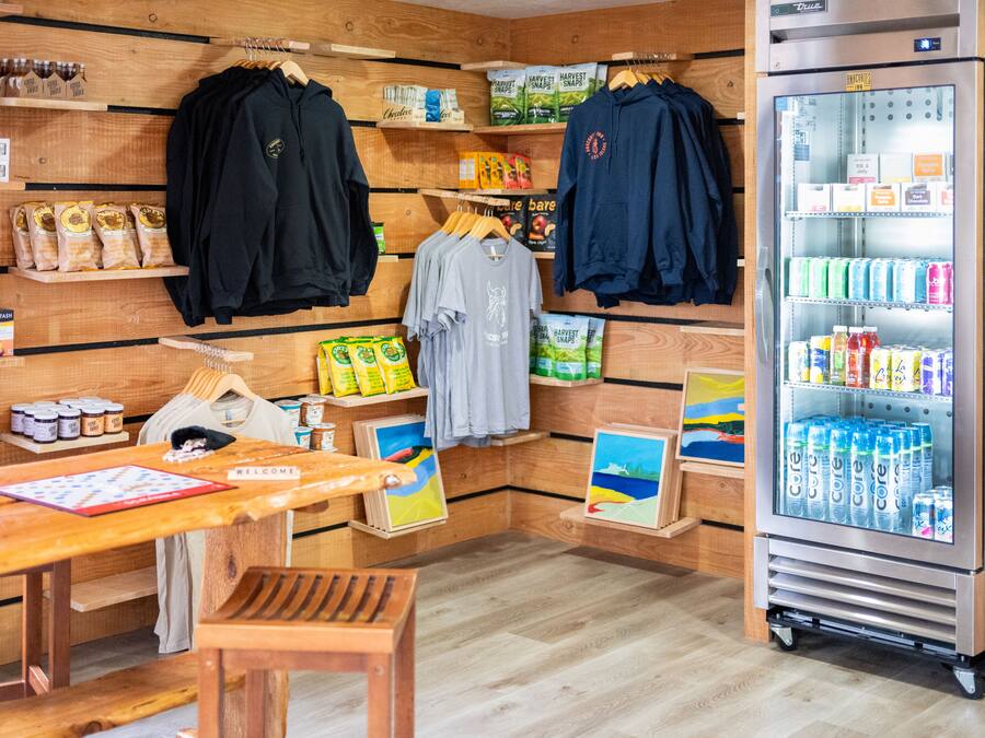 Una habitación con paredes de madera, una mesa de madera, un banco, un refrigerador lleno de bebidas y varios alimentos y ropa en estantes.
