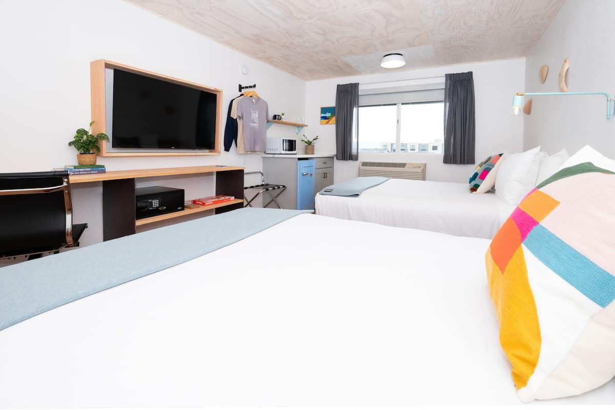 A hotel room with a bed, a desk with a computer, a TV, a small kitchenette, and a window with curtains.