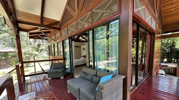 A covered outdoor area with a wooden deck, wicker furniture, and large glass doors leading to a bedroom. - Kenaki Lodge (Cahuita)