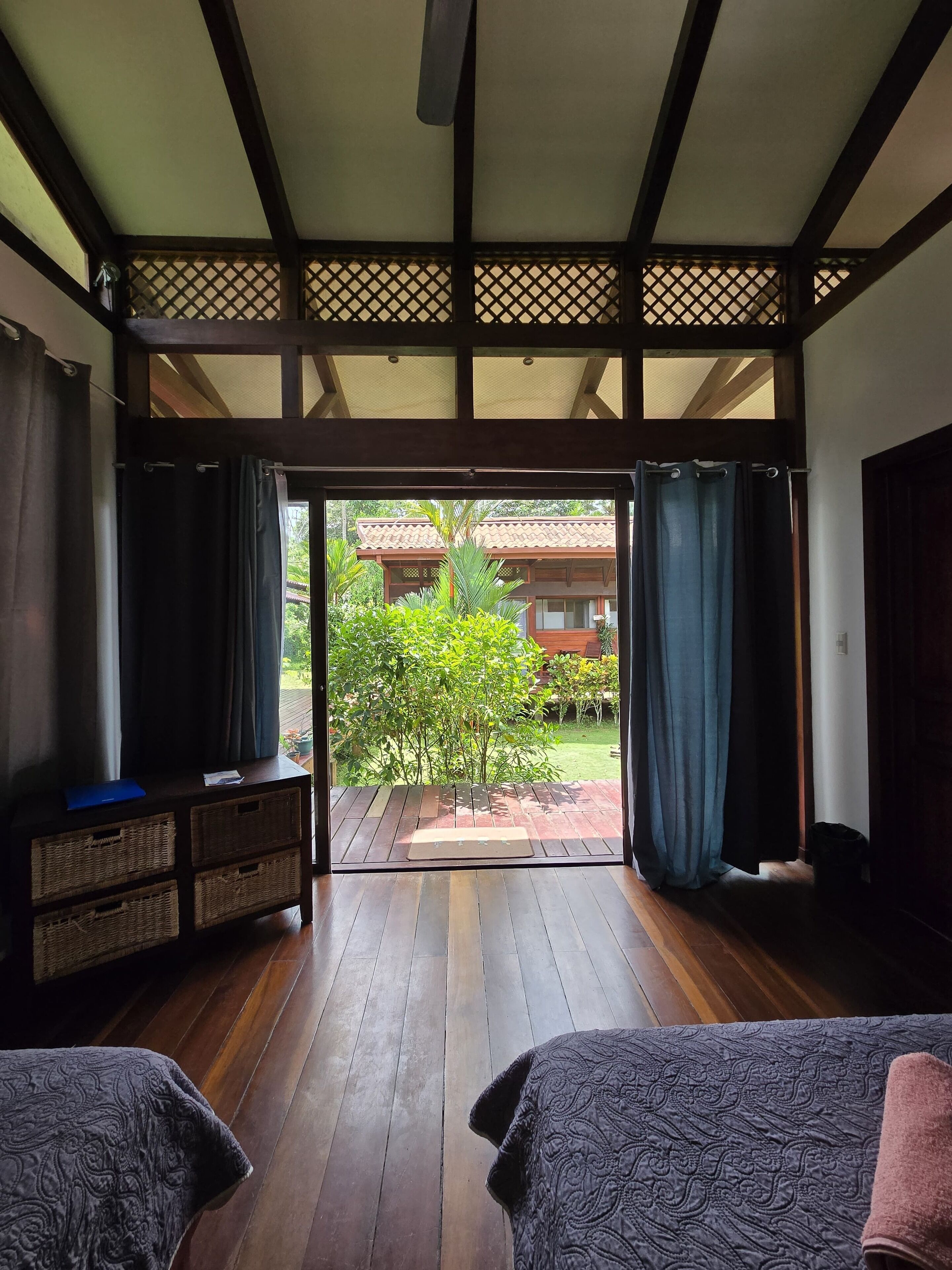 A room with a wooden floor, a bed with a patterned bedspread, a small table with a blue folder, and a view of a garden and building outside.