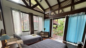 A room with two beds, a small table with a lamp, and a view of a garden through large windows. - Kenaki Lodge (Cahuita)